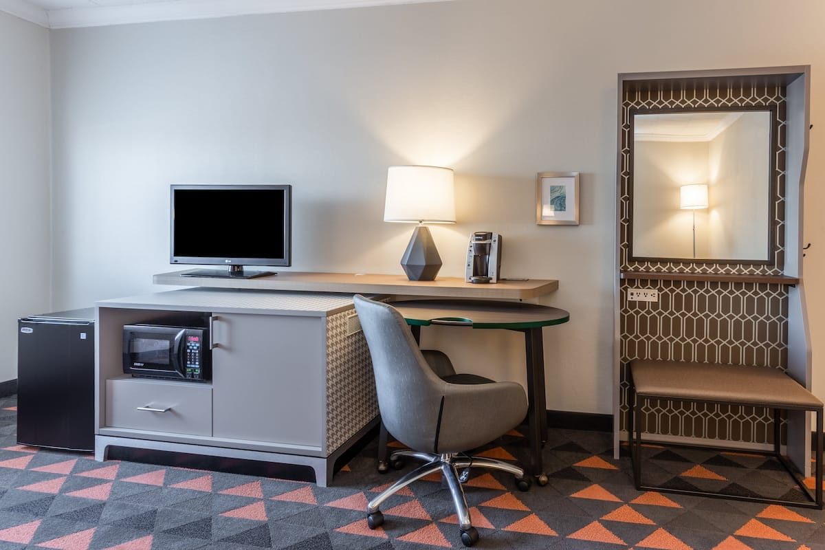 Ein Hotelzimmer mit Schreibtisch, Stuhl, kleinem Kühlschrank, Mikrowelle, Lampe, Spiegel und einem gemusterten Teppich.