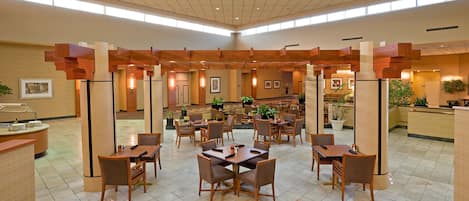 A spacious hotel lobby with multiple dining areas, ceiling fans, and natural light from large windows.