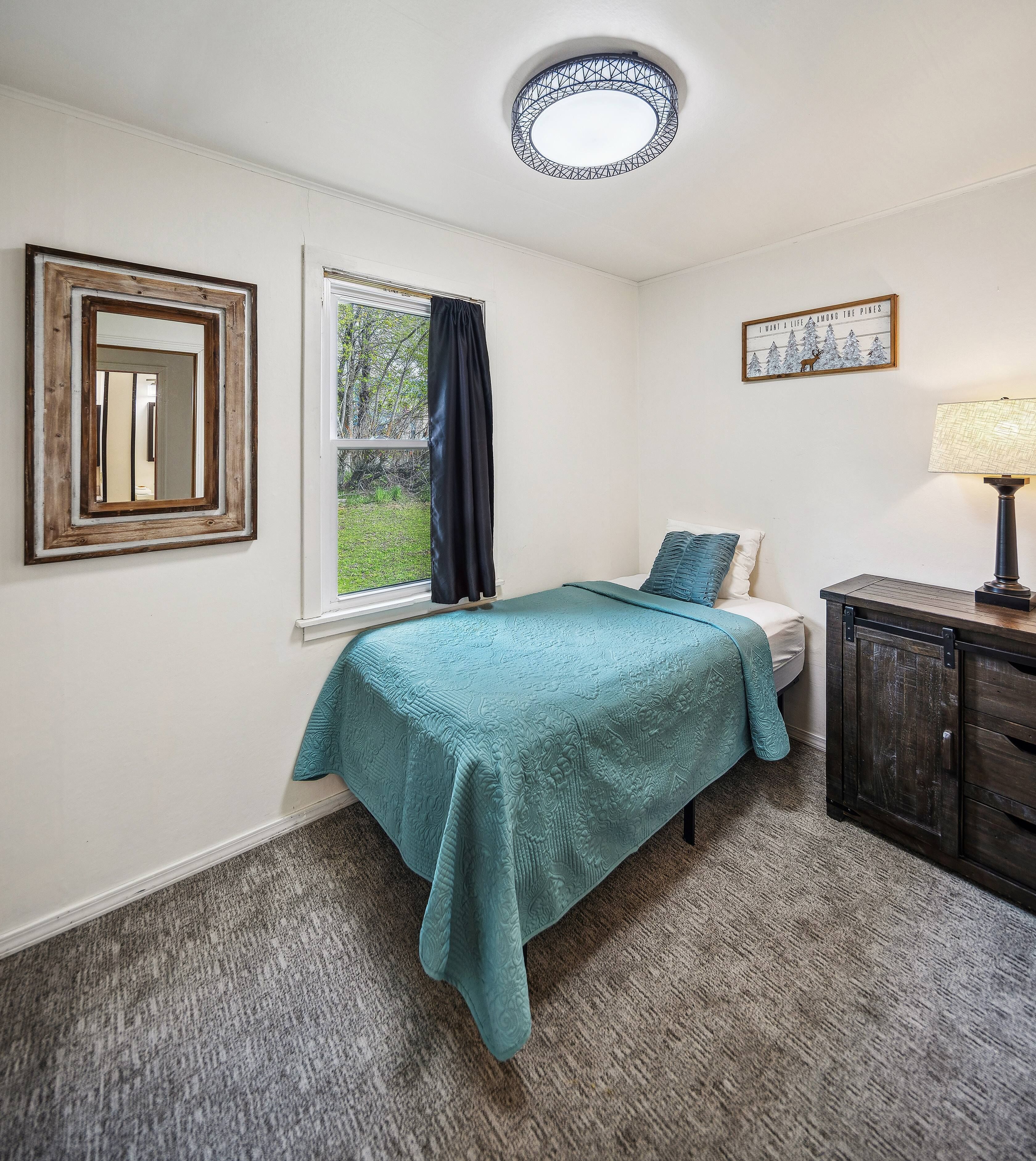Een slaapkamer met een bed, een raam met gordijnen, een lijst met afbeelding, een lamp en een kast.