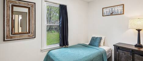 A bedroom with a bed, a window with curtains, a framed picture, a lamp, and a cabinet.