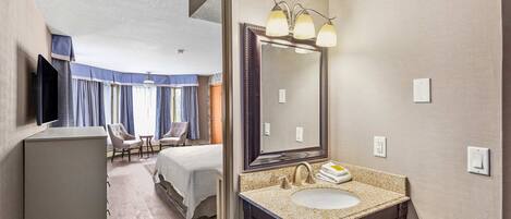 A hotel room with a large bed, a sitting area with a chair and table, a bathroom with a sink and mirror, and a television on the wall.