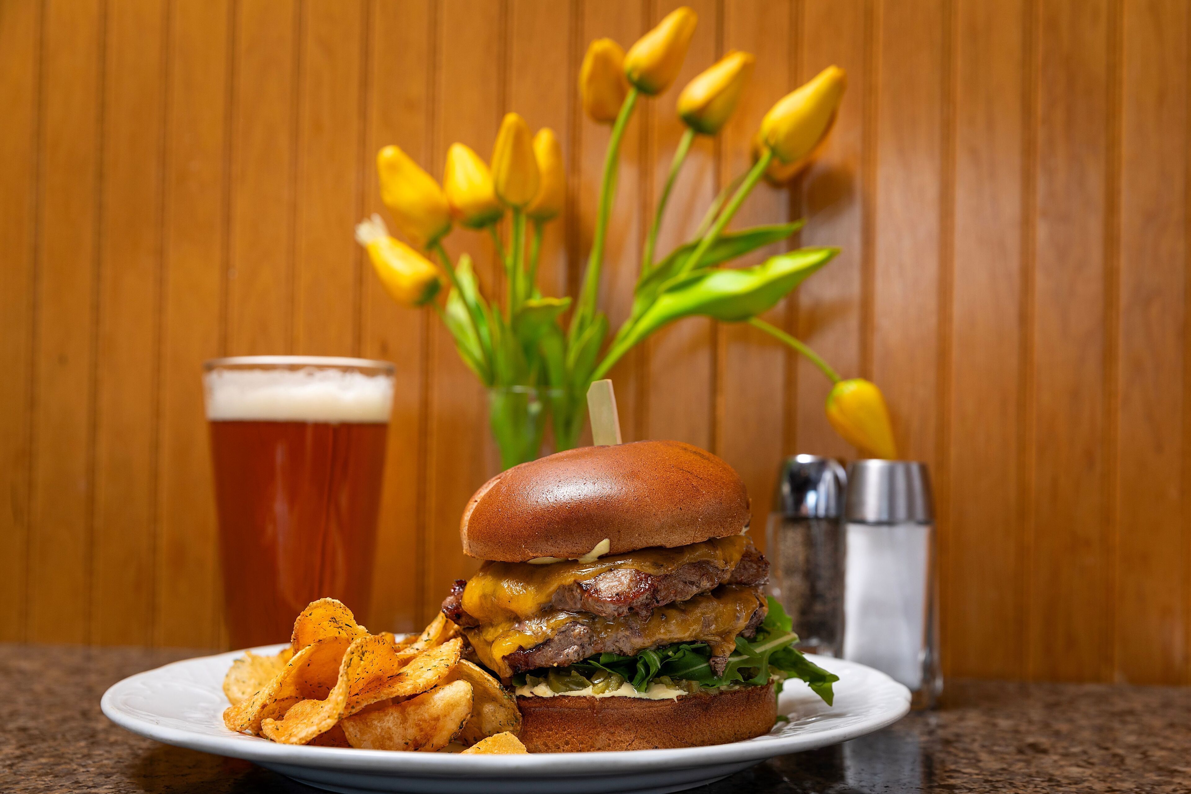 A burger with fries and a glass of beer on a plate, with a bouquet of yellow flowers in the background.