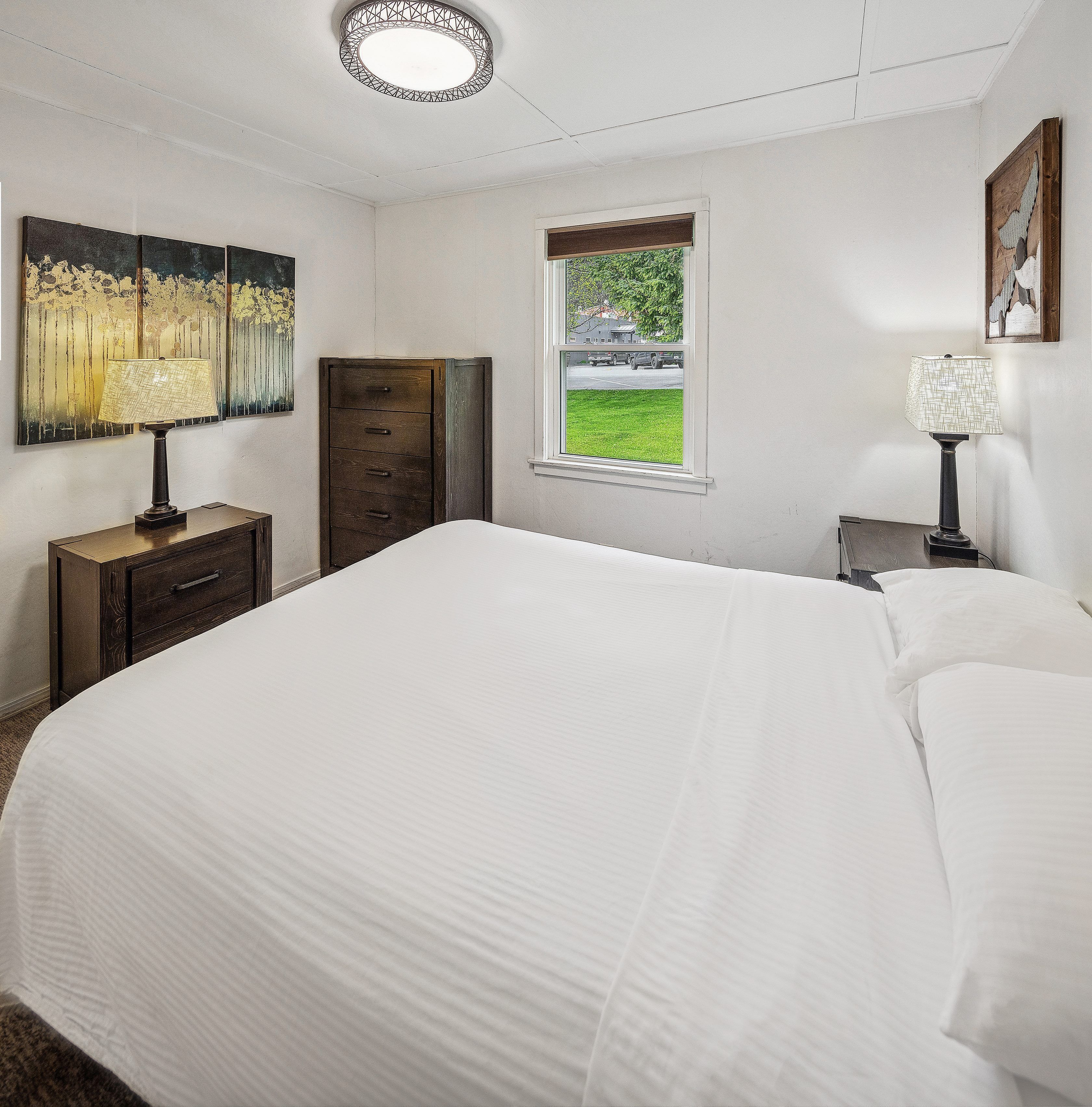 A bedroom with a large bed, a dresser, a window with a view of the outdoors, and a ceiling light fixture.
