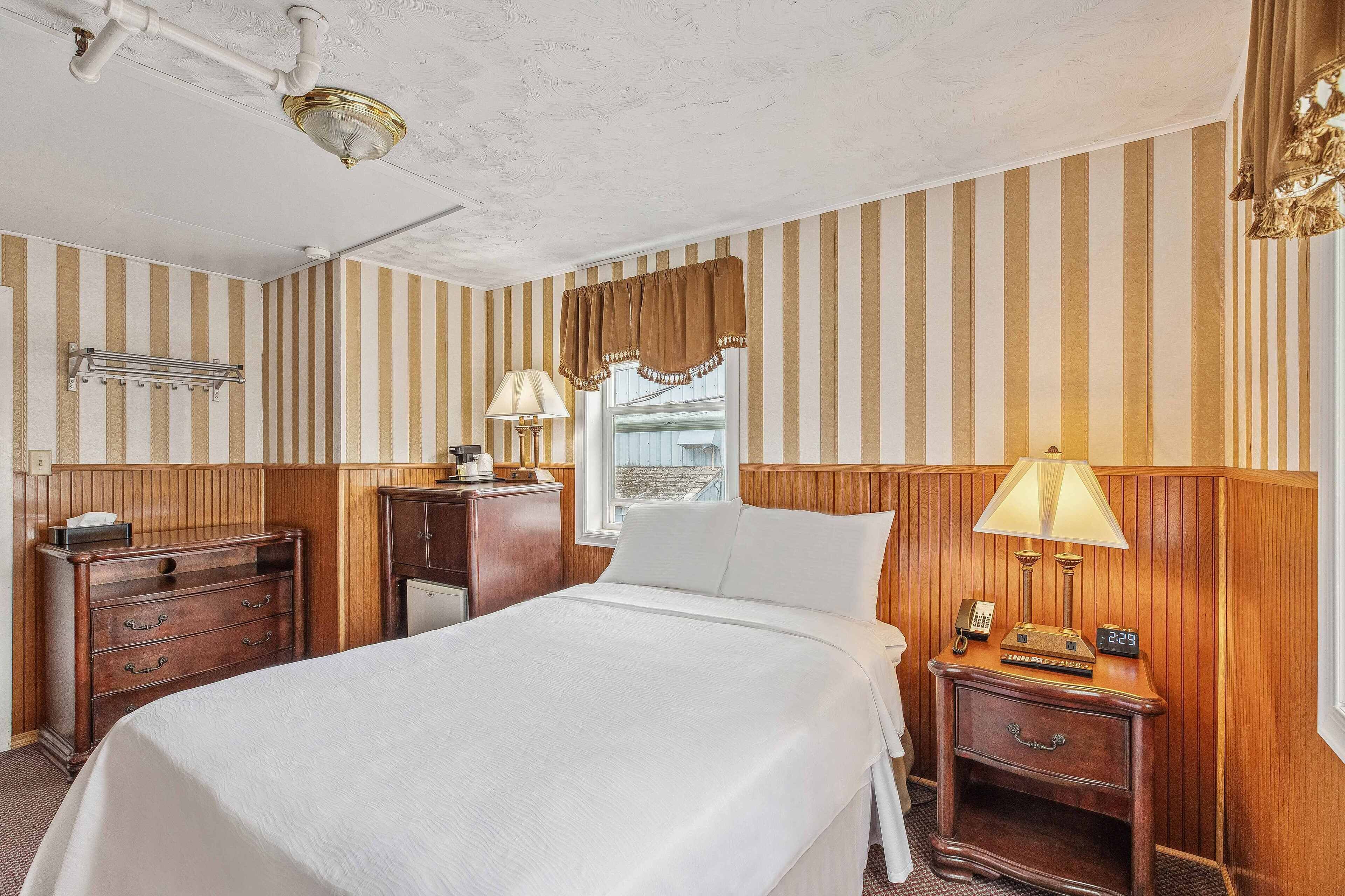 A hotel room with a large bed, wooden headboard, a nightstand with a lamp, a dresser, and a window with striped curtains.