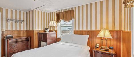 Habitación de hotel con una cama grande, cabecera de madera, mesita de noche con lámpara, cómoda y ventana con cortinas a rayas.