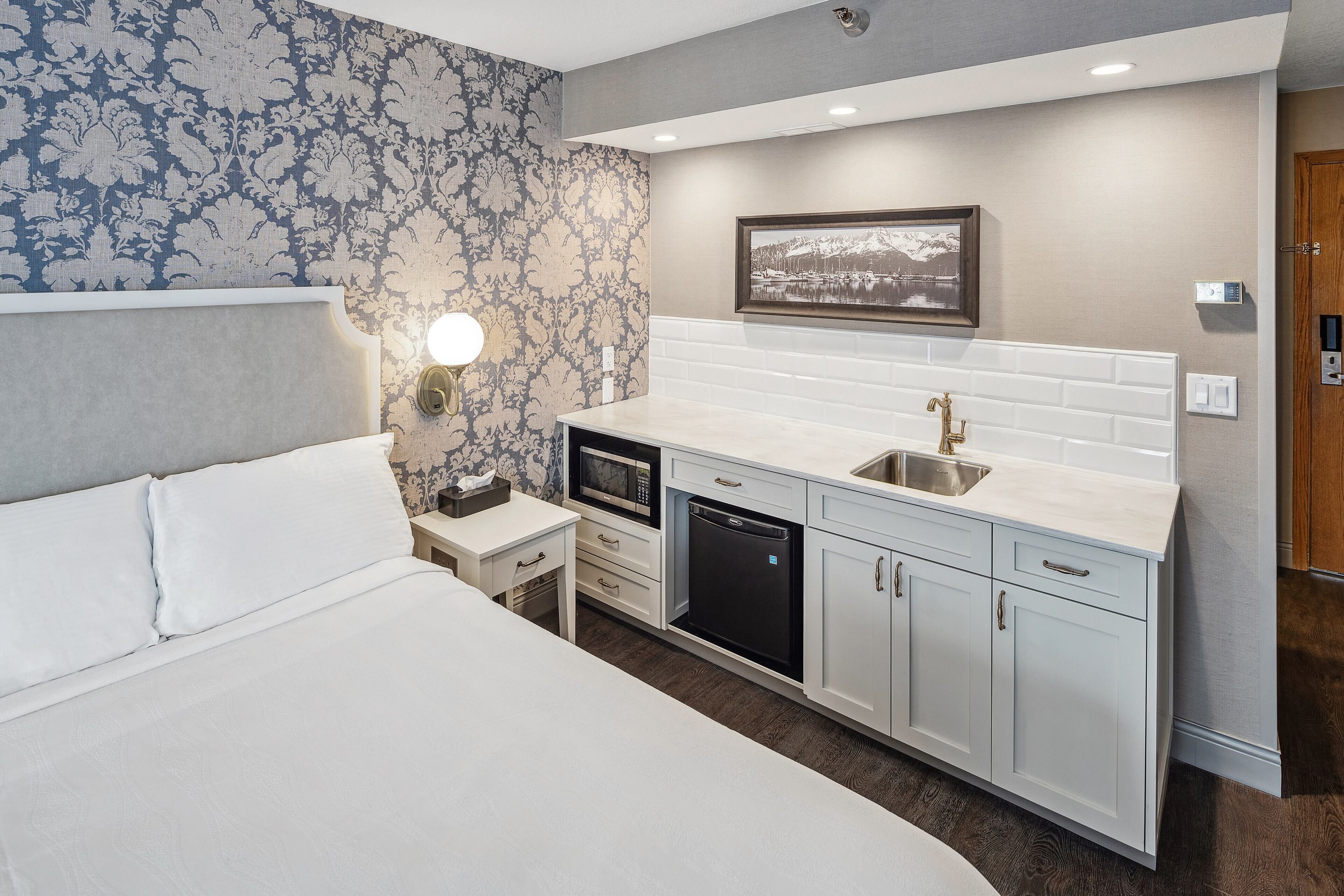 A hotel room with a bed, a bedside table with a lamp, a microwave, a dishwasher, and a sink.