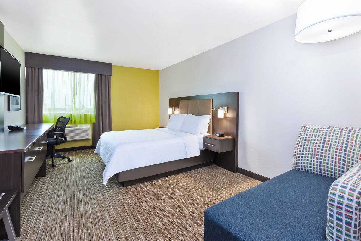 Une chambre d’hôtel avec un lit, un bureau, une chaise, une télévision et une fenêtre avec des rideaux.
