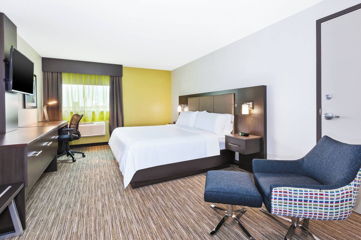 Une chambre d’hôtel avec un lit, un bureau, une chaise, une télévision et une fenêtre avec des rideaux.