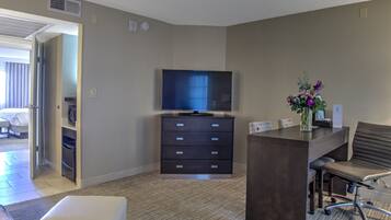 A hotel room with a flat-screen TV, a desk with a chair, a white ottoman, and a vase with flowers.