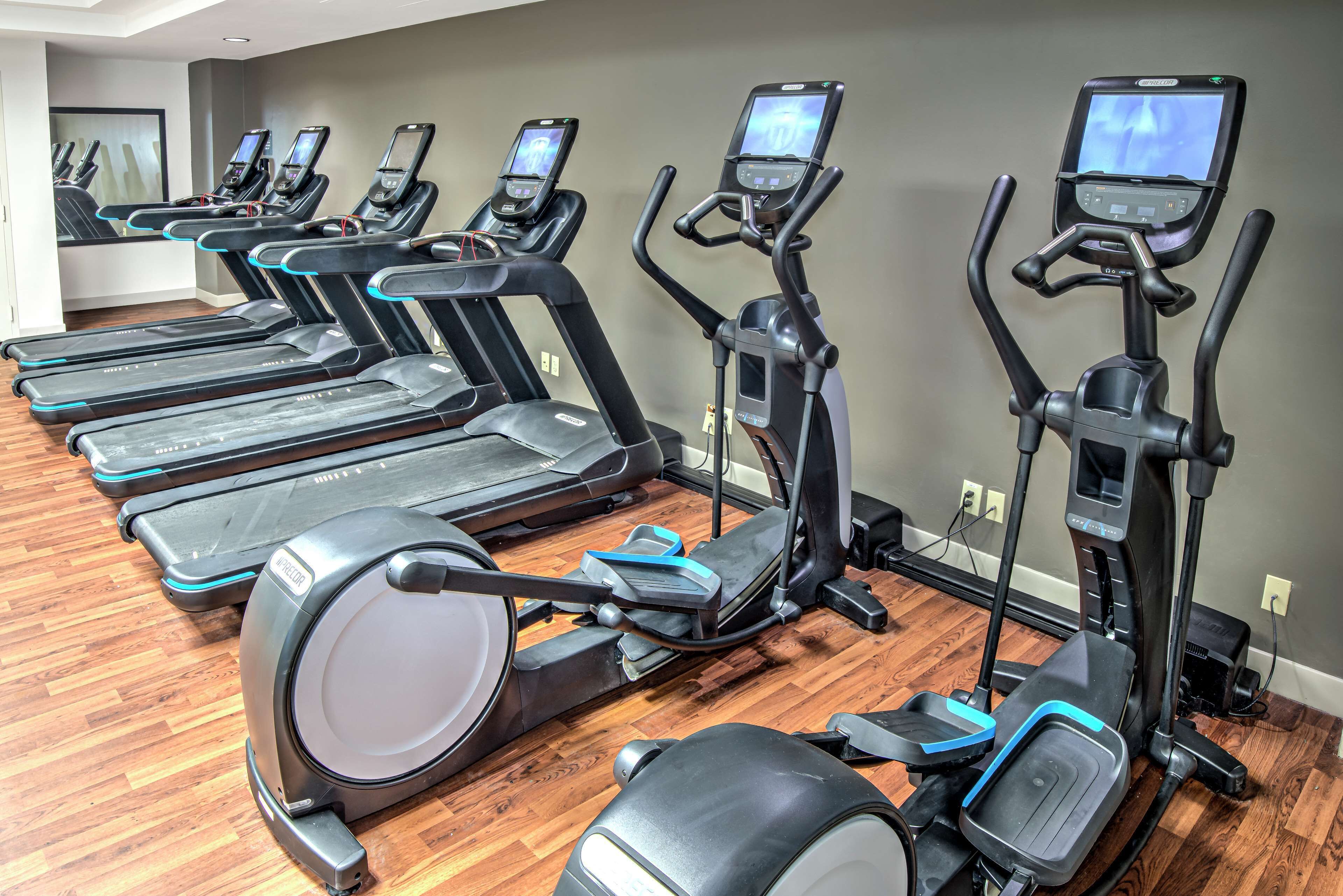 A row of stationary bikes and treadmills in a gym with wooden flooring and gray walls.