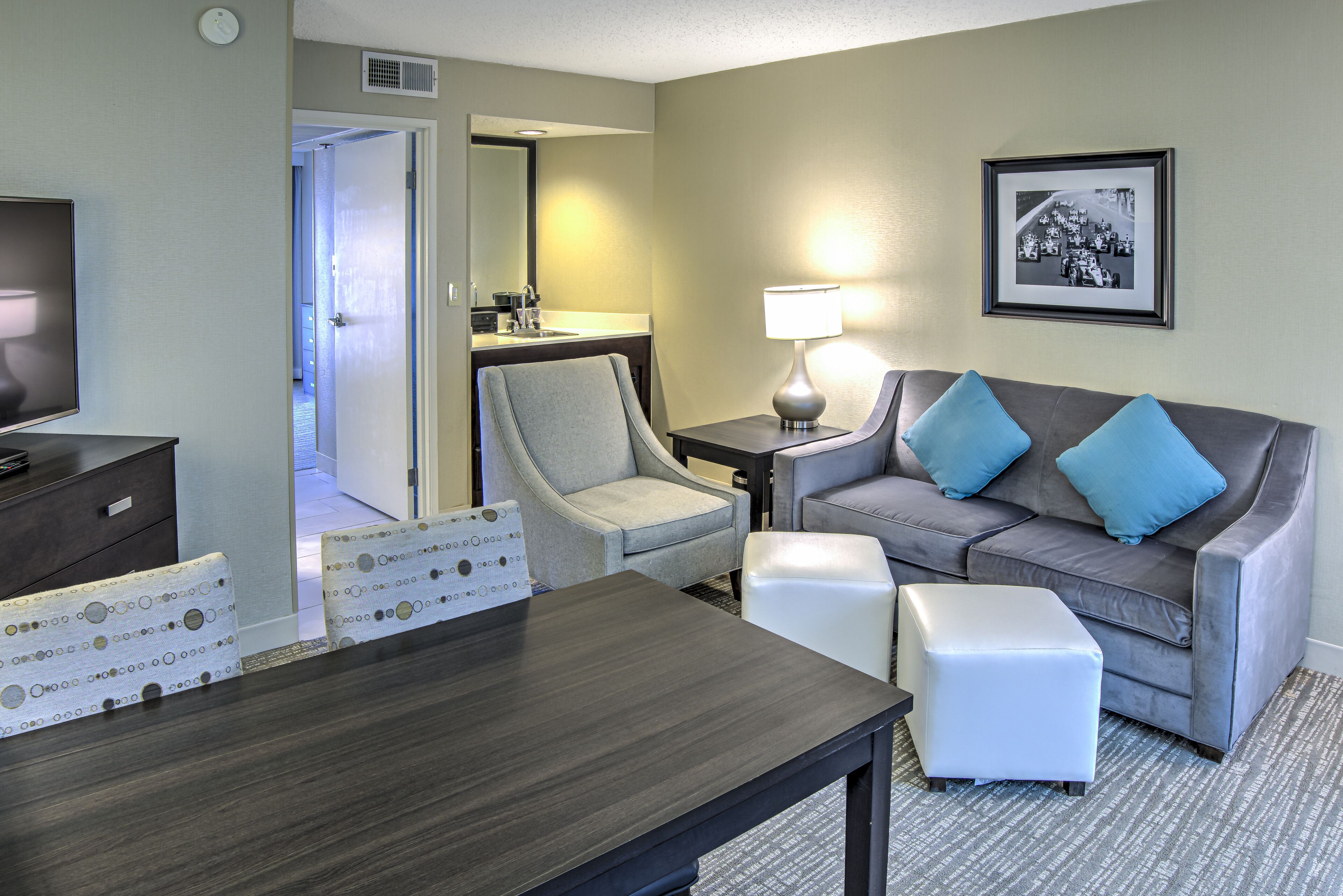 A hotel room with a dark wood table, a grey sofa with blue cushions, a white armchair, a small white ottoman, a framed photo on the wall, and a television on a dark wooden dresser.
