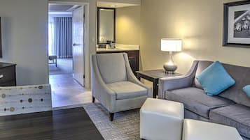 Habitación de hotel con un sillón gris, un sofá gris con un cojín azul, un otoman blanco, una mesita de noche con una lámpara, un espejo y un cuadro en la pared.