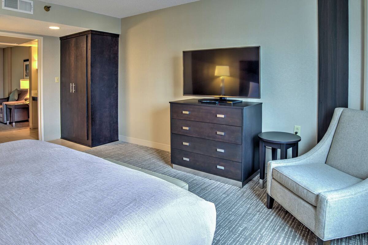 A hotel room with a large bed, a dark wooden dresser, a flat-screen TV, a small round table, and a grey armchair.