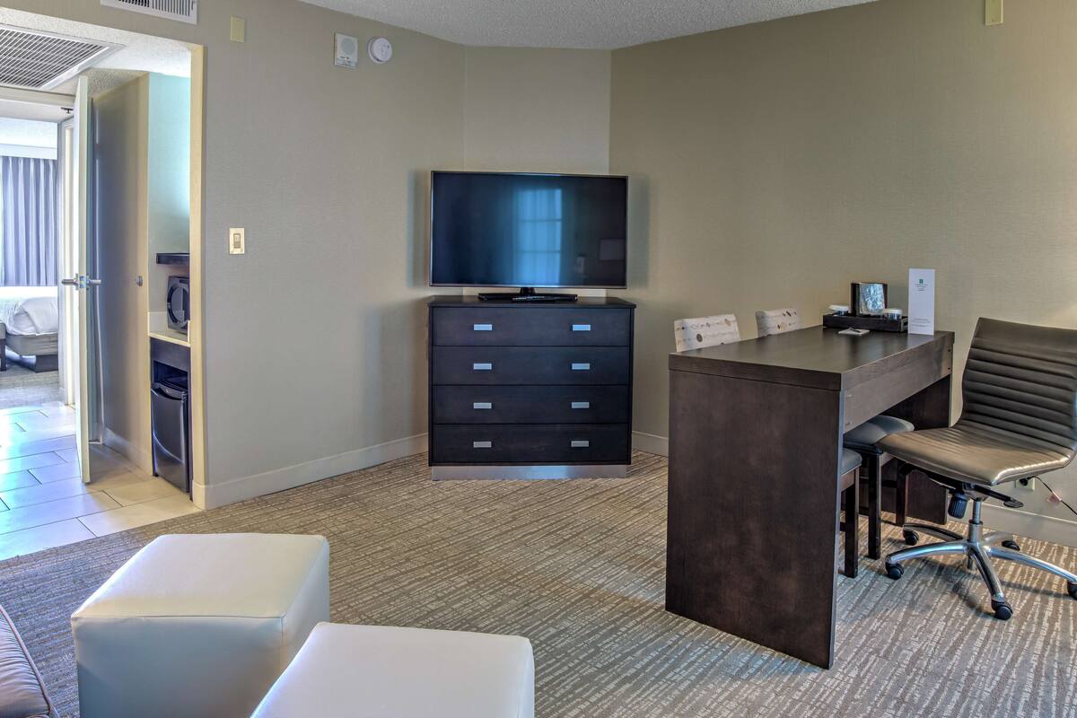 A hotel room with a flat-screen TV, a desk with a chair, a minibar, and a large window.