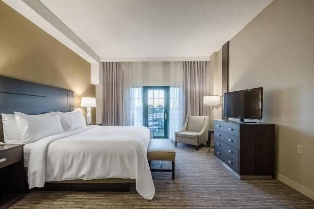 A hotel room with a bed, a TV, a chair, a dresser, and a view of the outdoors through a window.
