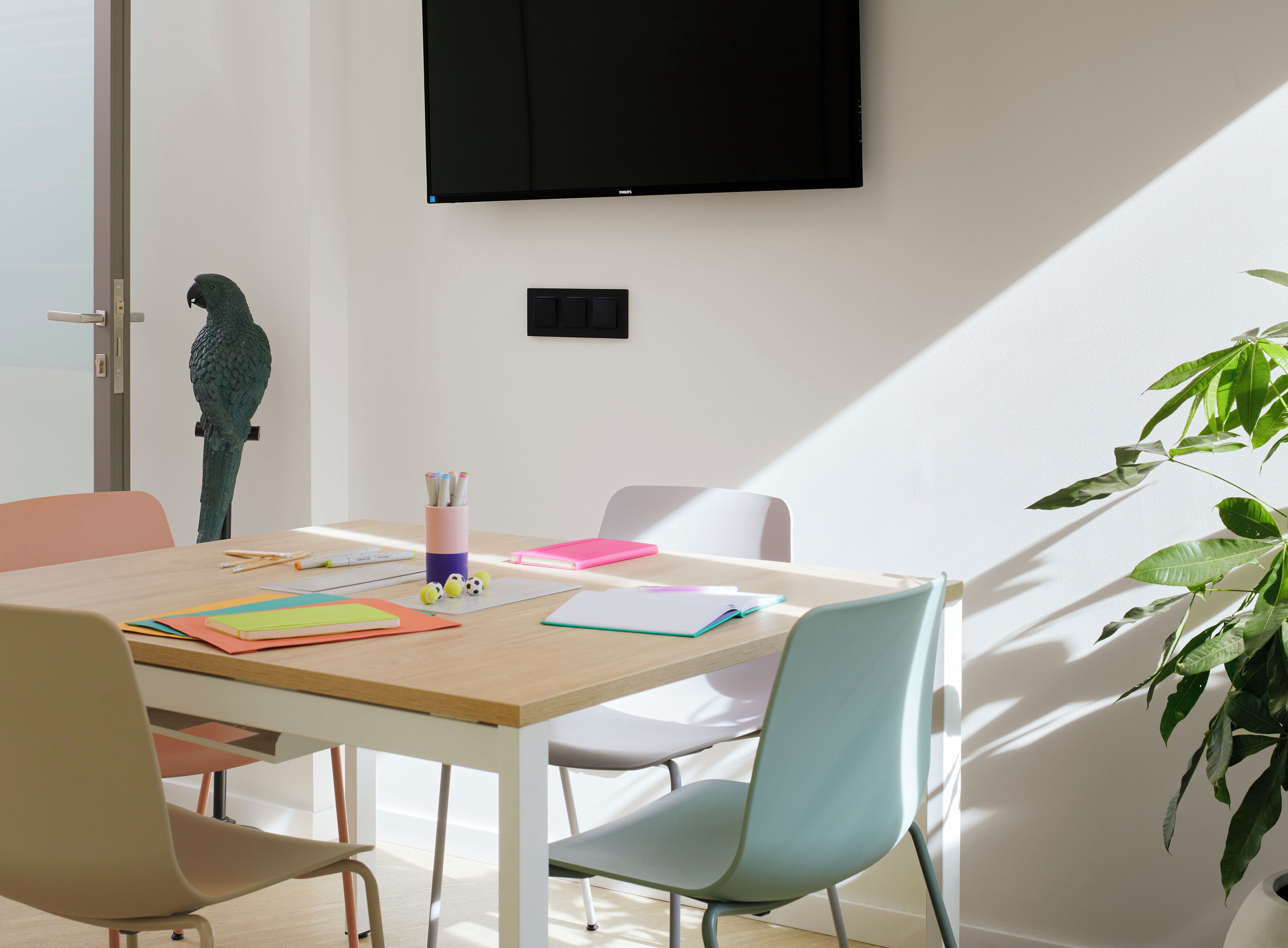 Una habitación moderna con una mesa de madera, sillas coloridas, un televisor montado y un ventanal con luz natural.