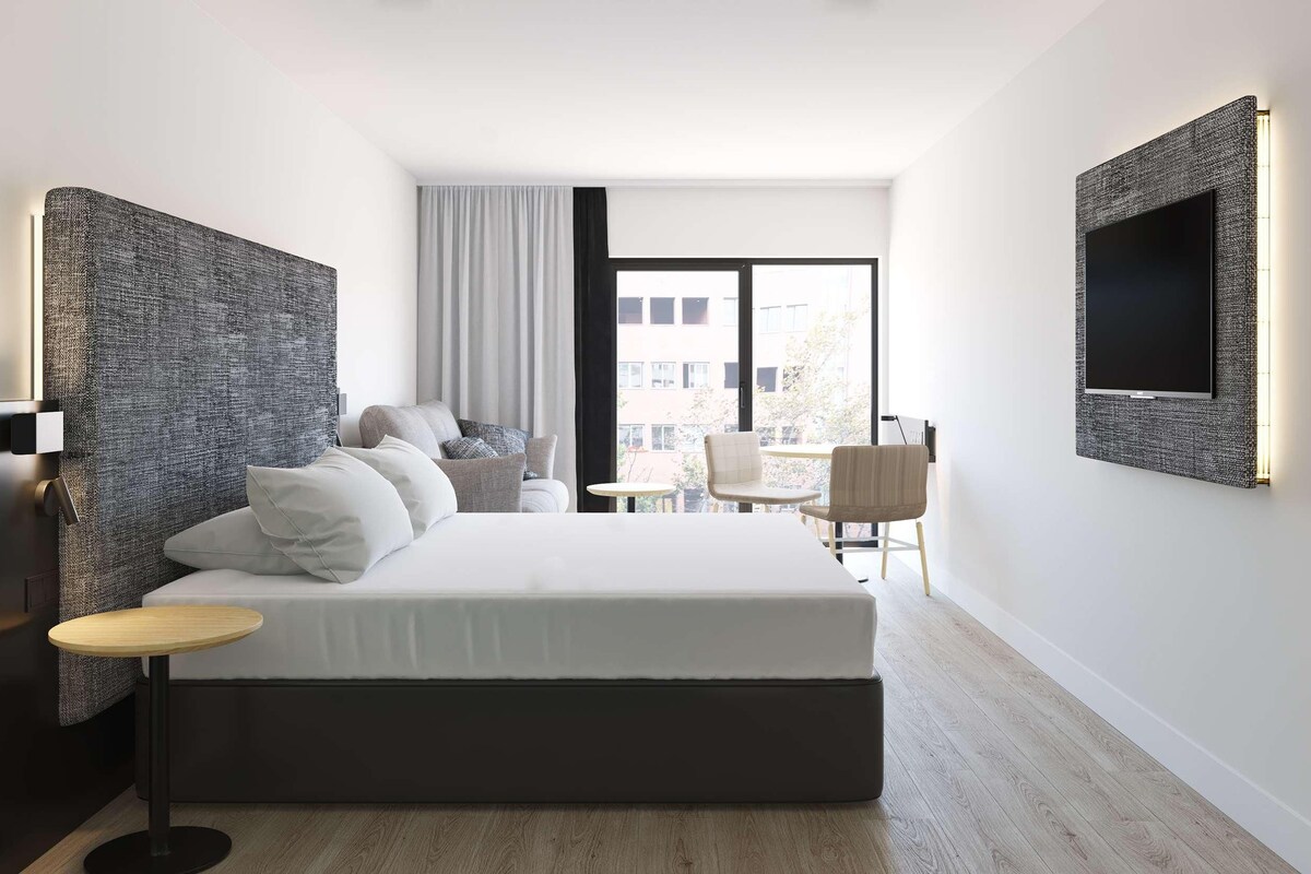 Ein modernes Hotelzimmer mit einem großen Bett, einem kleinen runden Tisch, einem Stuhl, einem an der Wand befestigten Fernseher und Blick auf die Stadt durch das Fenster.