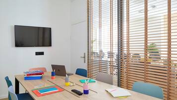 Uma sala de conferências com uma mesa grande, cadeiras, um laptop, livros e uma TV na parede.