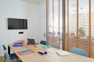 Uma sala de conferências com uma mesa grande, cadeiras, um laptop, livros e uma TV na parede.