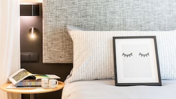 Una cama bien hecha con un cuadro enmarco de ojos, una mesita con libros y una taza, y una lámpara fijada a la pared.