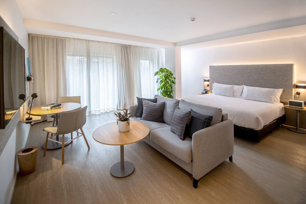 Una habitación de hotel moderna con cama, sofá, mesa redonda y una planta en maceta.