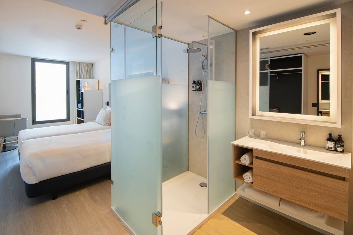 Una habitación de hotel moderna con ducha envuelta en vidrio, cama con ropa blanca, tocador de madera con espejo y un amplio ventanal que proporciona luz natural.