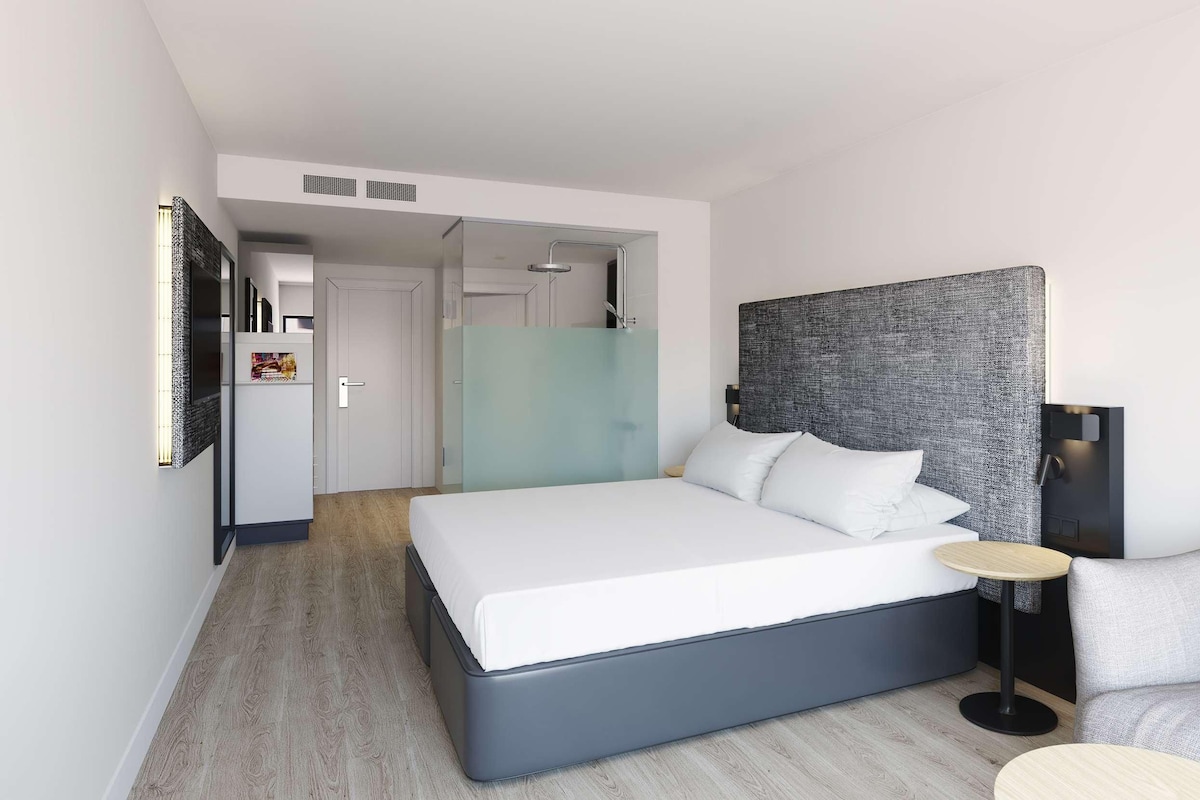 Une chambre d’hôtel moderne avec un grand lit, un canapé gris, une petite table ronde et un téléviseur fixé au mur.