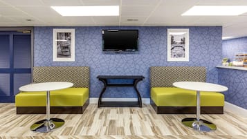 A reception area with a blue patterned wall, a flat-screen TV mounted above a black console table, and two yellow cushioned seating areas with round tables.