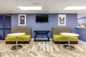 A reception area with a blue patterned wall, a flat-screen TV mounted above a black console table, and two yellow cushioned seating areas with round tables.