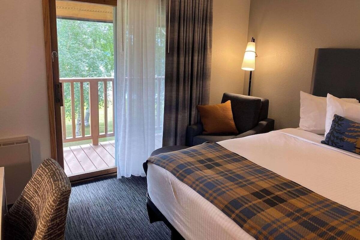 A hotel room with a bed, a chair, a lamp, and a view of greenery through the window.