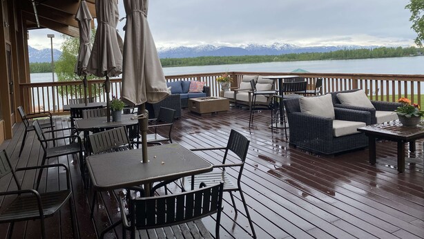 A wooden deck with outdoor furniture, including tables and chairs, overlooking a lake with mountains in the background. - Best Western Lake Lucille Inn (Wasilla)