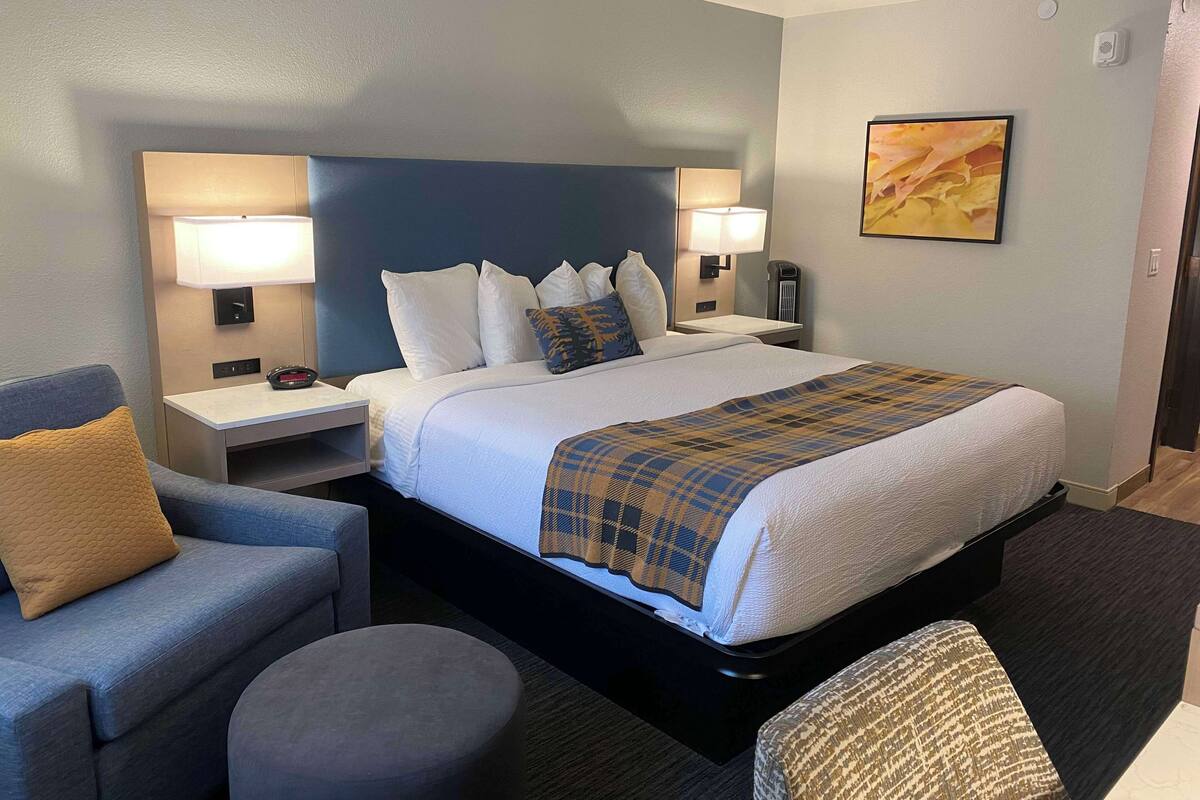 A hotel room with a large bed, two bedside tables, a chair, a round ottoman, and a painting on the wall.