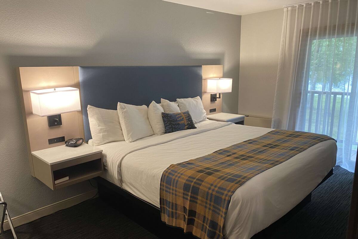A hotel room with a large bed, two bedside tables, a nightstand, a lamp, and a window with curtains.