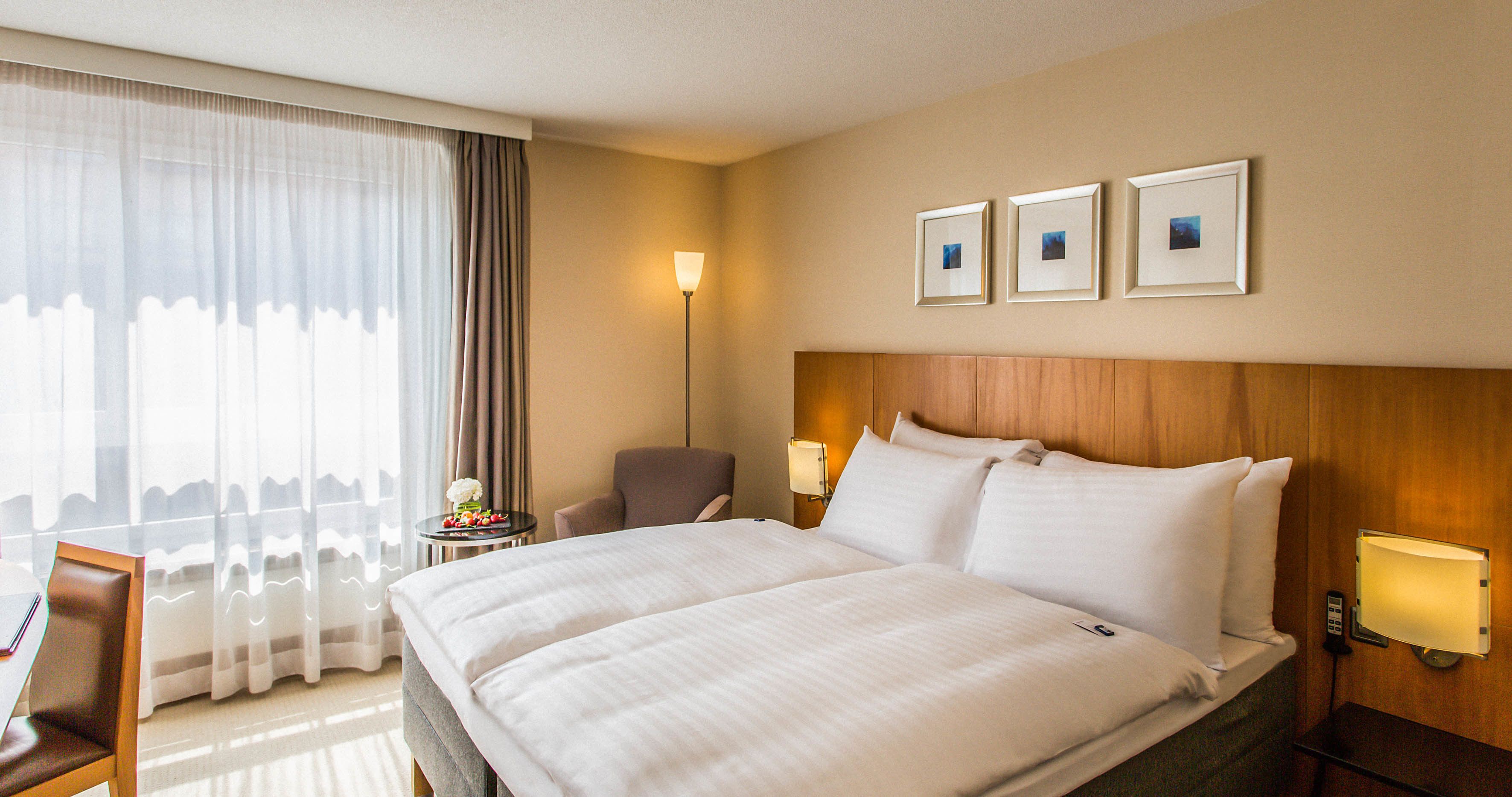 Ein Hotelzimmer mit einem großen Bett, einem Sessel, einem kleinen Tisch mit Lampe sowie drei gerahmten Bildern an der Wand.