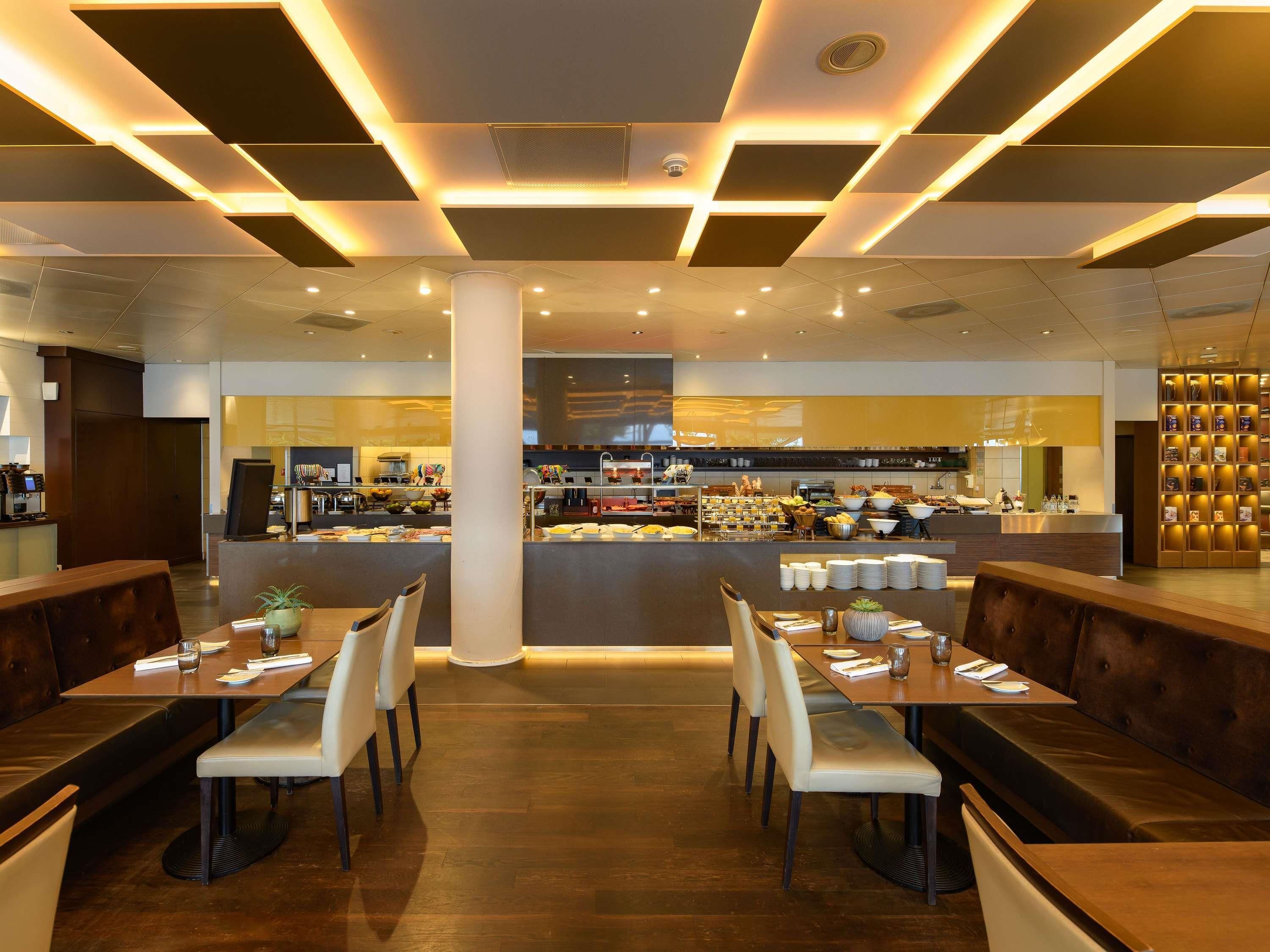 A modern restaurant with a central counter, multiple seating areas, and a visible kitchen in the background.
