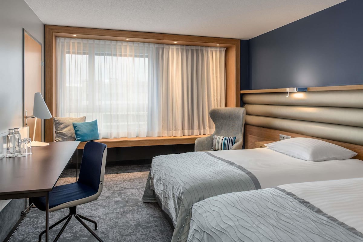 Een hotelkamer met een bed, een bureau met stoel, een raam met gordijnen en een hoofdeinde.