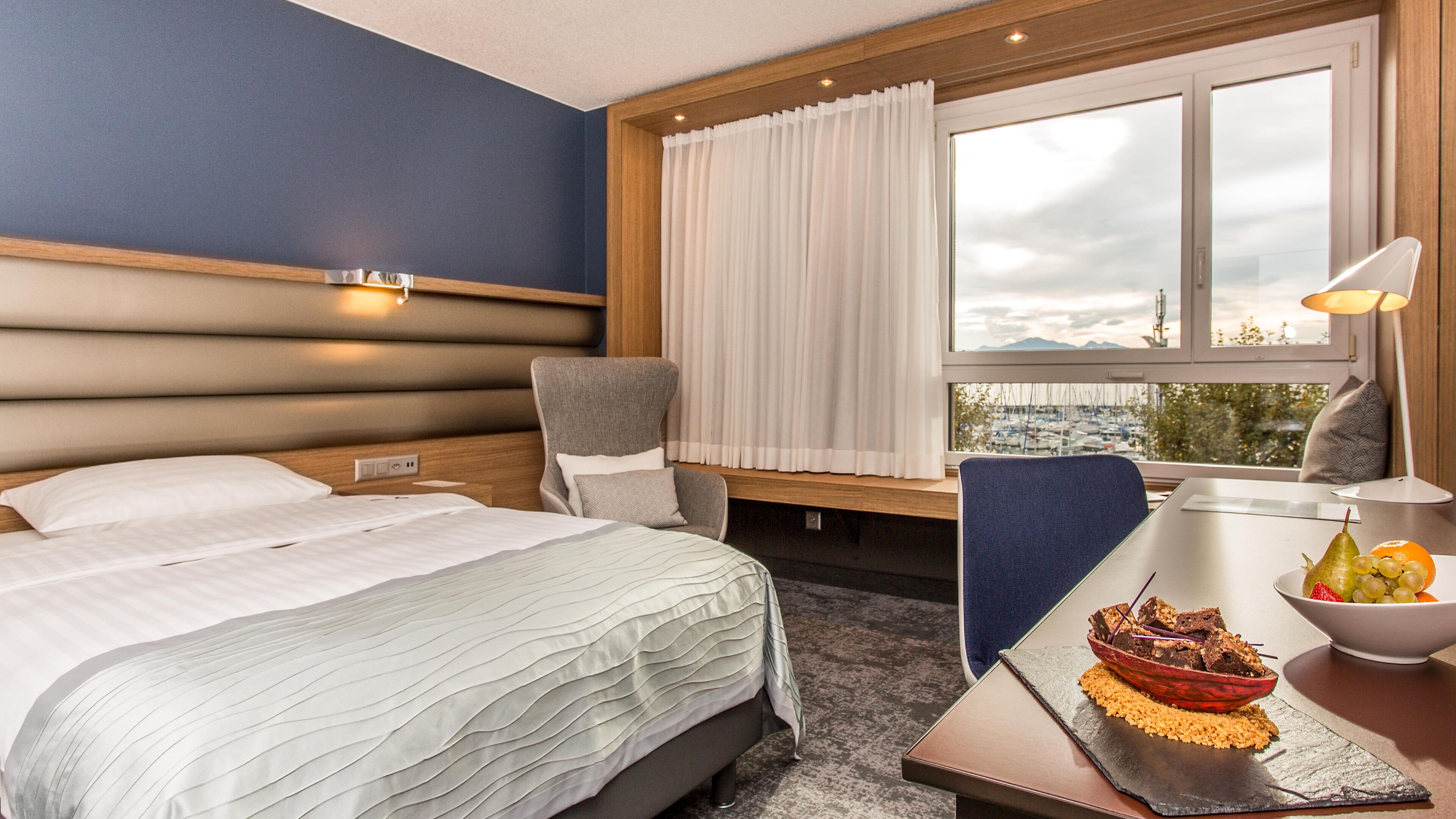 Hotelkamer met een bed, bureau met een schaal fruit en een schaal eten, een stoel, een raam met gordijnen en uitzicht op de stad.