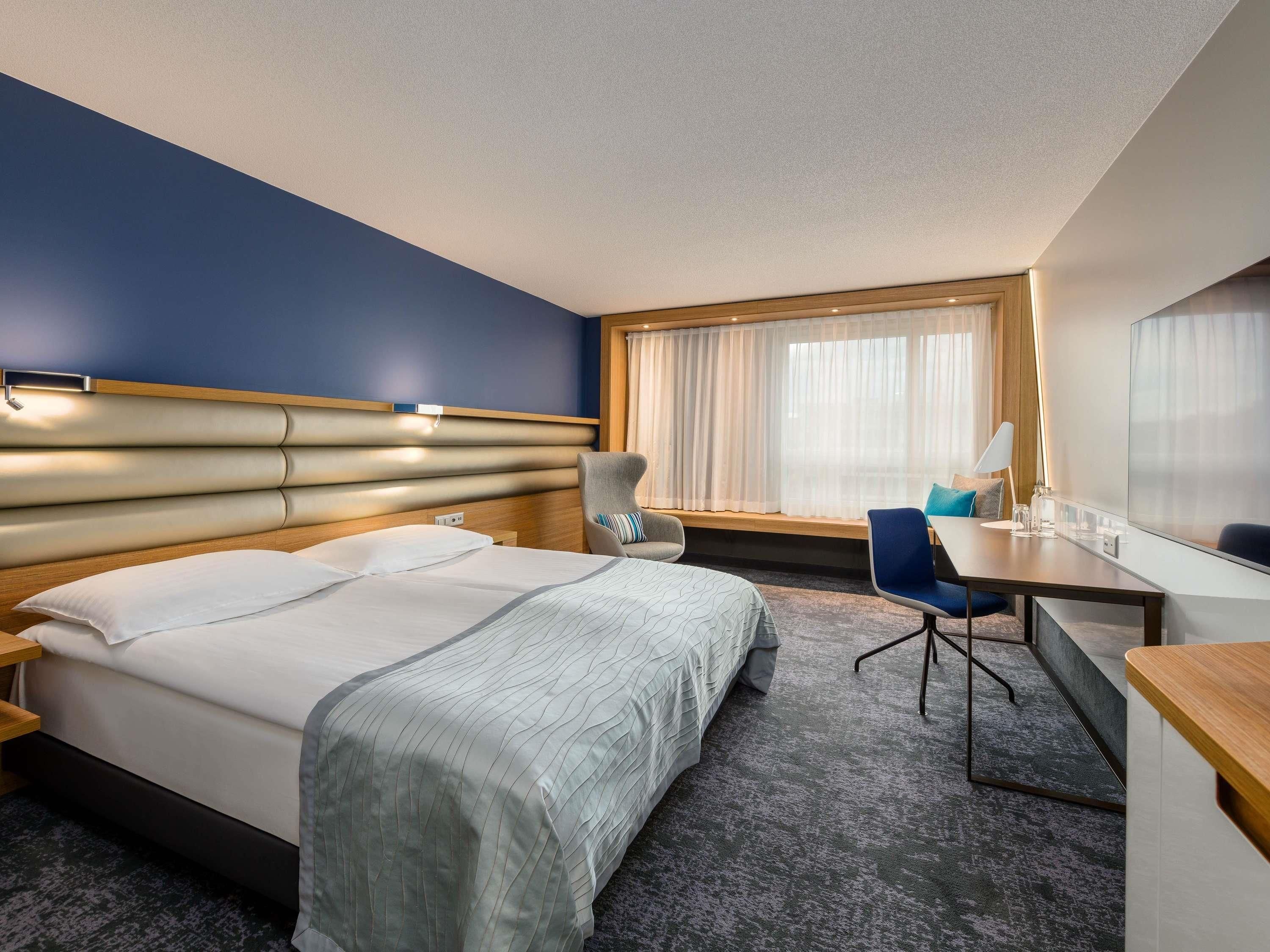 Ein Hotelzimmer mit einem großen Bett, einem Schreibtisch, einem Stuhl und einem Fenster mit Vorhängen.