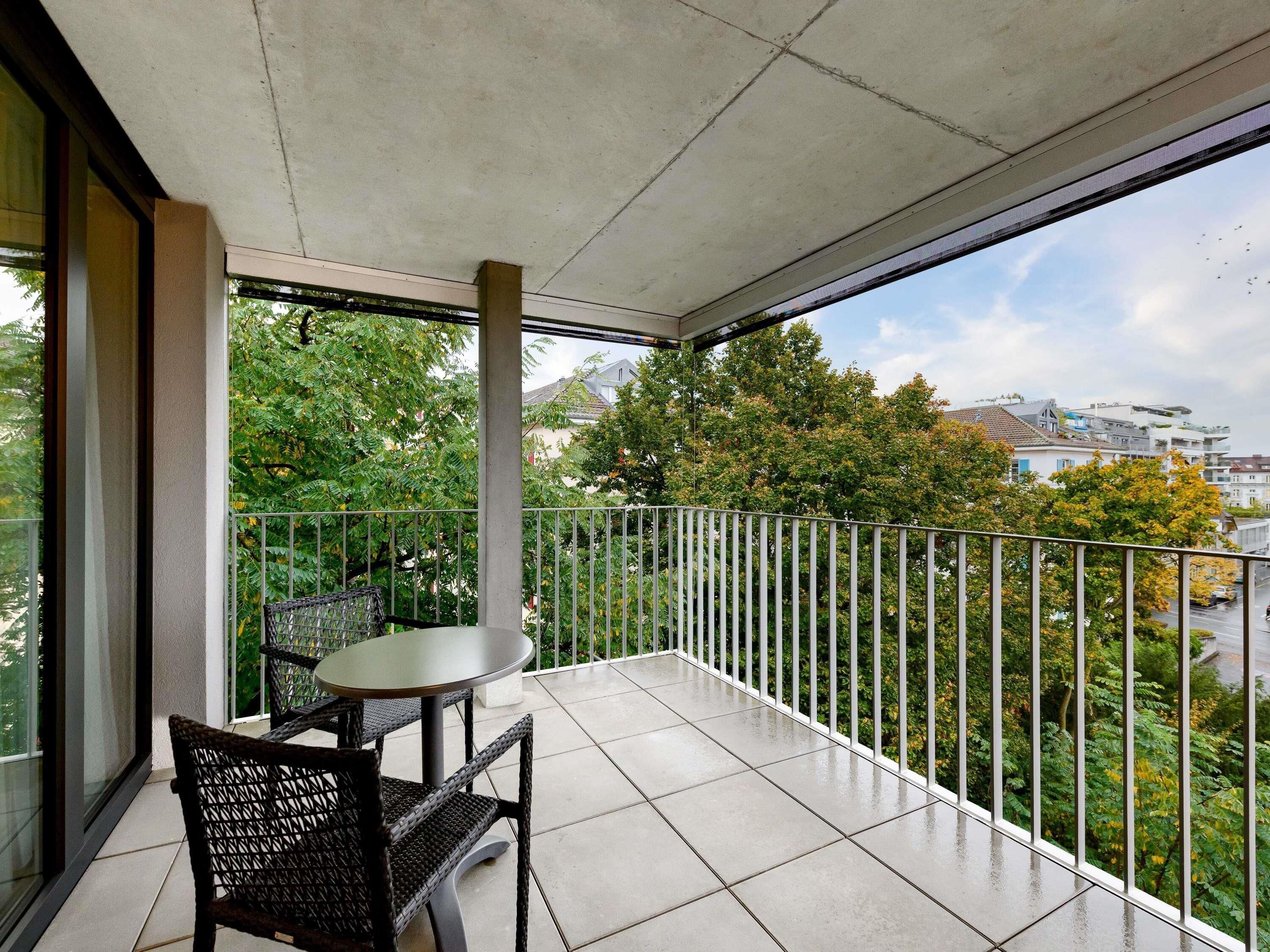 A balcony with a table and chairs, a view of trees and buildings, and a metal railing.
