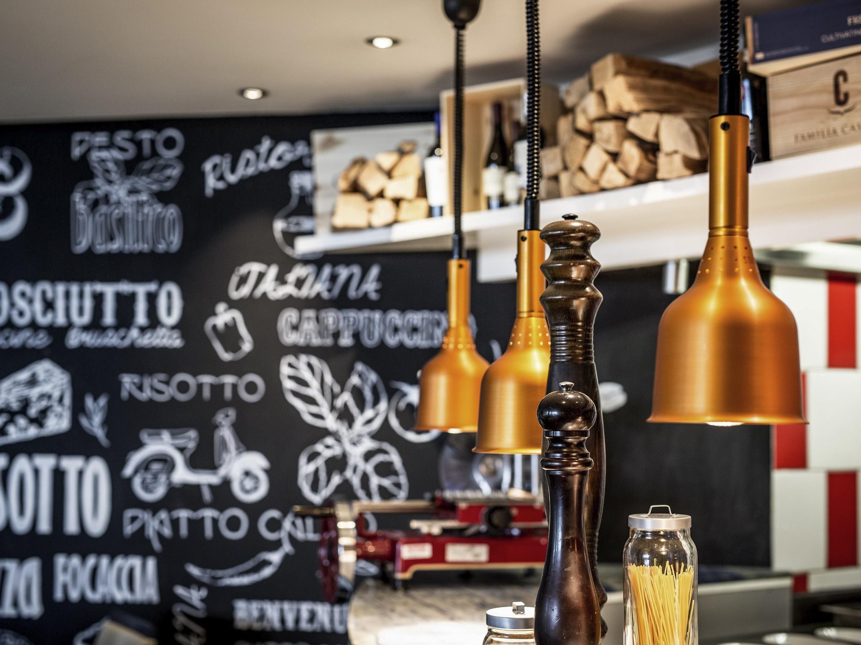 A kitchen with hanging copper-colored pendant lights, a glass jar with pasta, and a wall with various food-related logos.