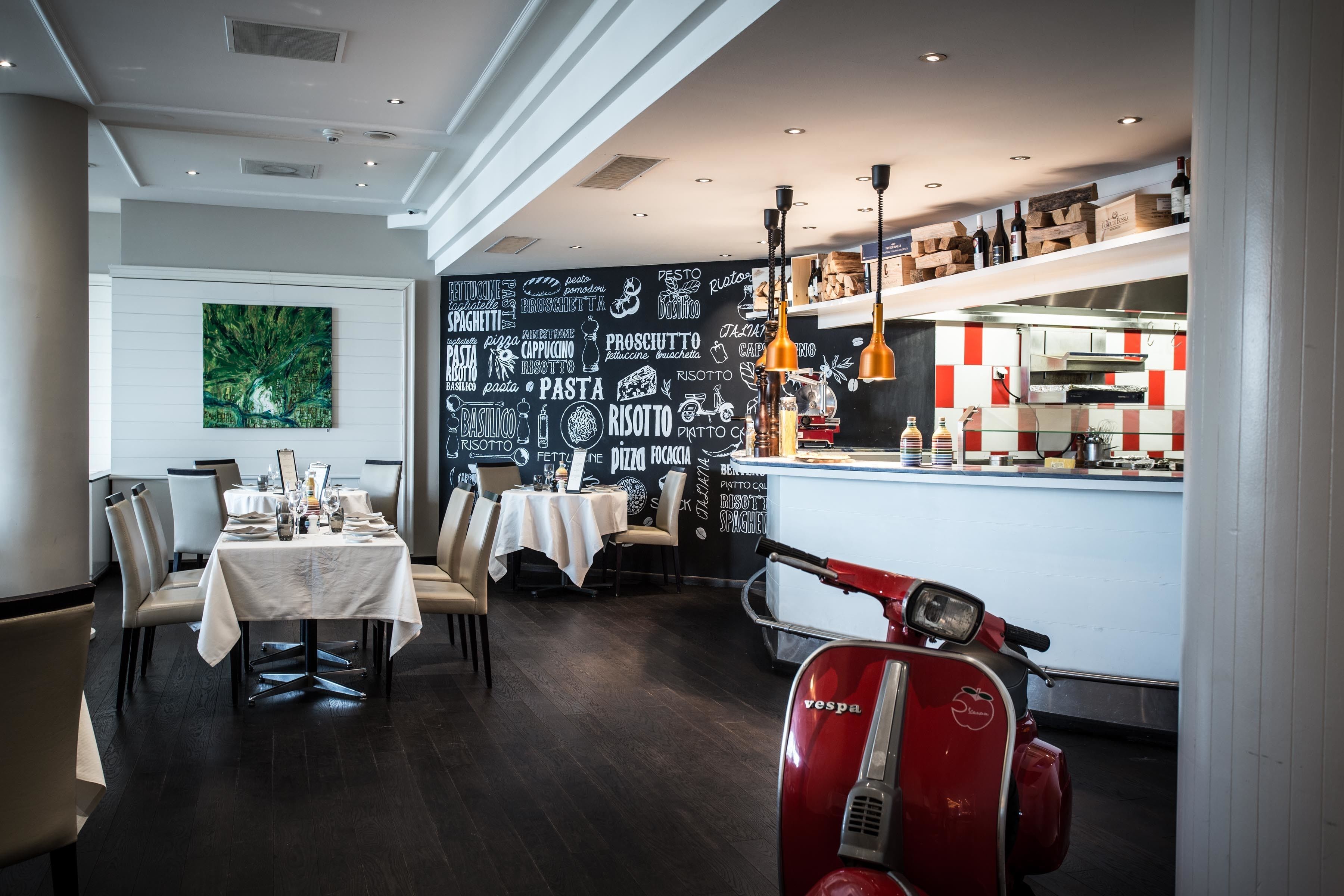 Un restaurante con una pared de pizarra decorada con palabras relacionadas a la comida, una zona de comedor con mesas y sillas, y una Vespa roja.