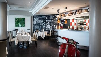 Un restaurante con una pared de pizarra decorada con palabras relacionadas a la comida, una zona de comedor con mesas y sillas, y una Vespa roja.