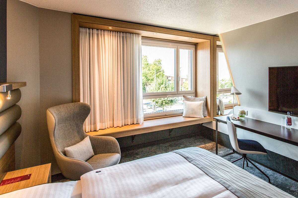Una camera d'albergo con un letto, una scrivania, una sedia, una TV e una finestra con tende.