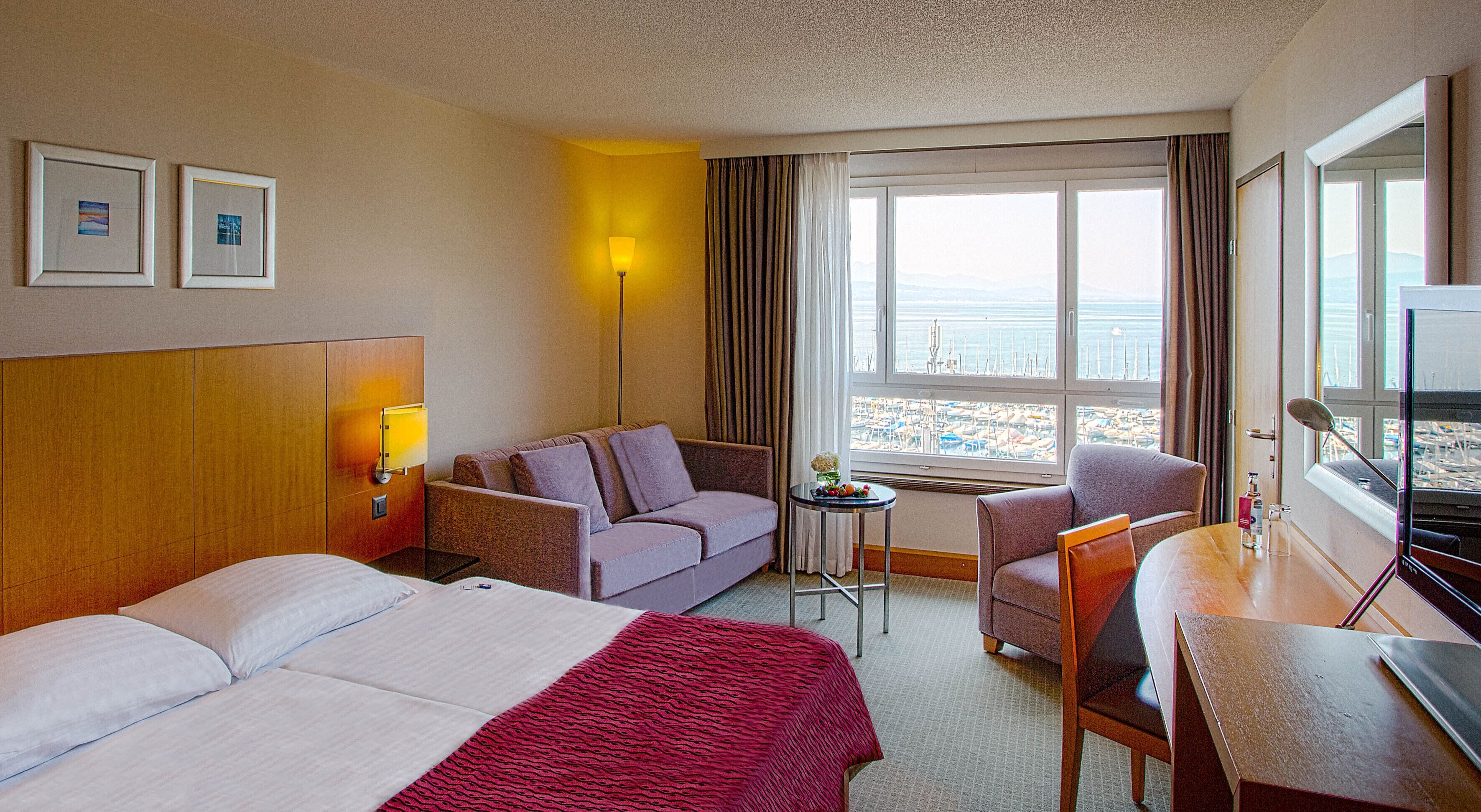 Ein Hotelzimmer mit Bett, einer Couch, einem Schreibtisch und einem Stuhl. An der Wand hängen zwei gerahmte Bilder, eine Lampe und ein Fenster mit Blick auf den Strand.