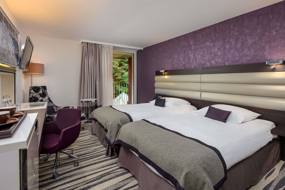 Een hotelkamer met twee bedden, een bureau, een stoel en uitzicht op groen buiten.