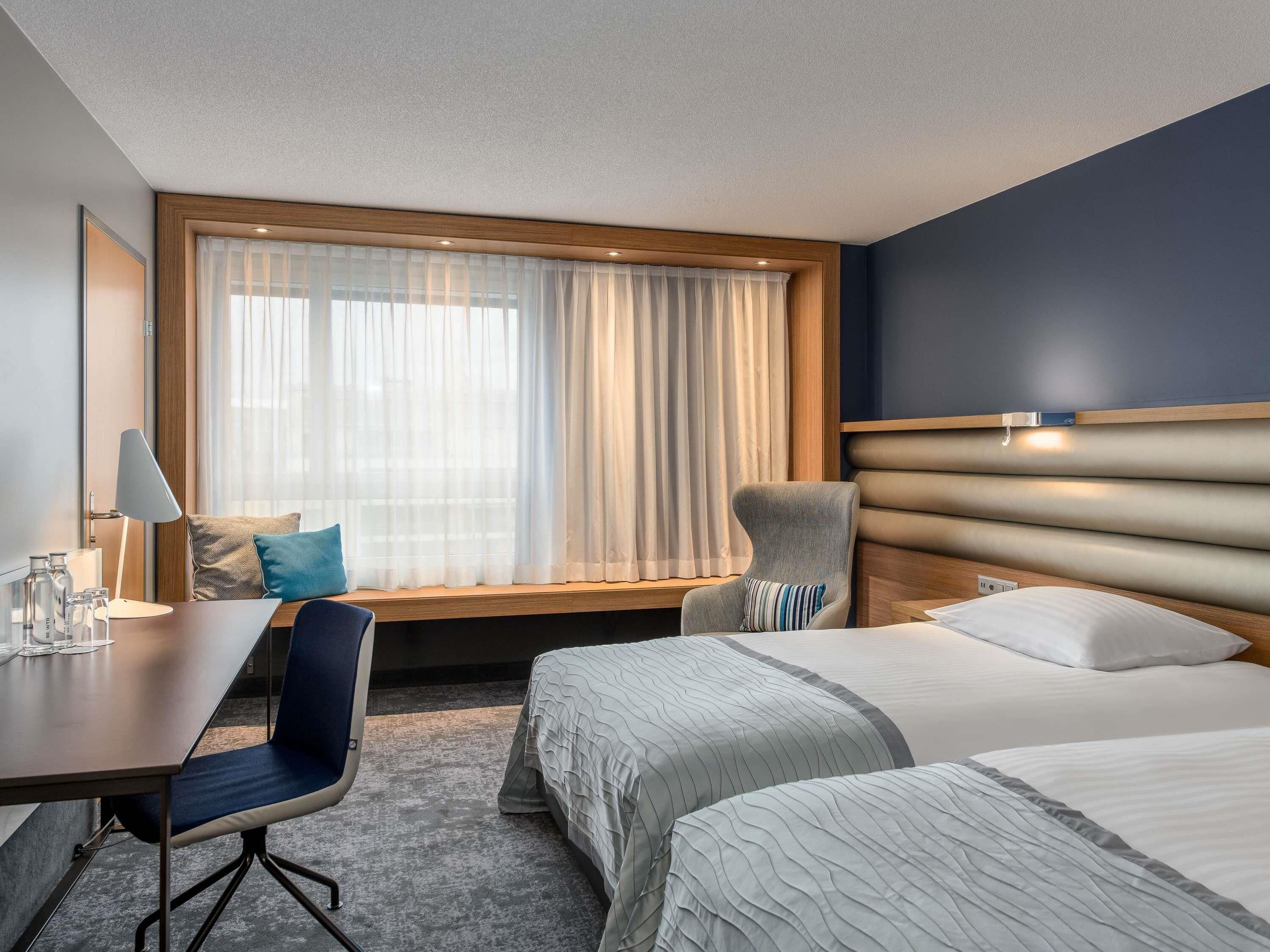 A hotel room with a bed, a desk with a chair, a window with curtains, and a headboard.