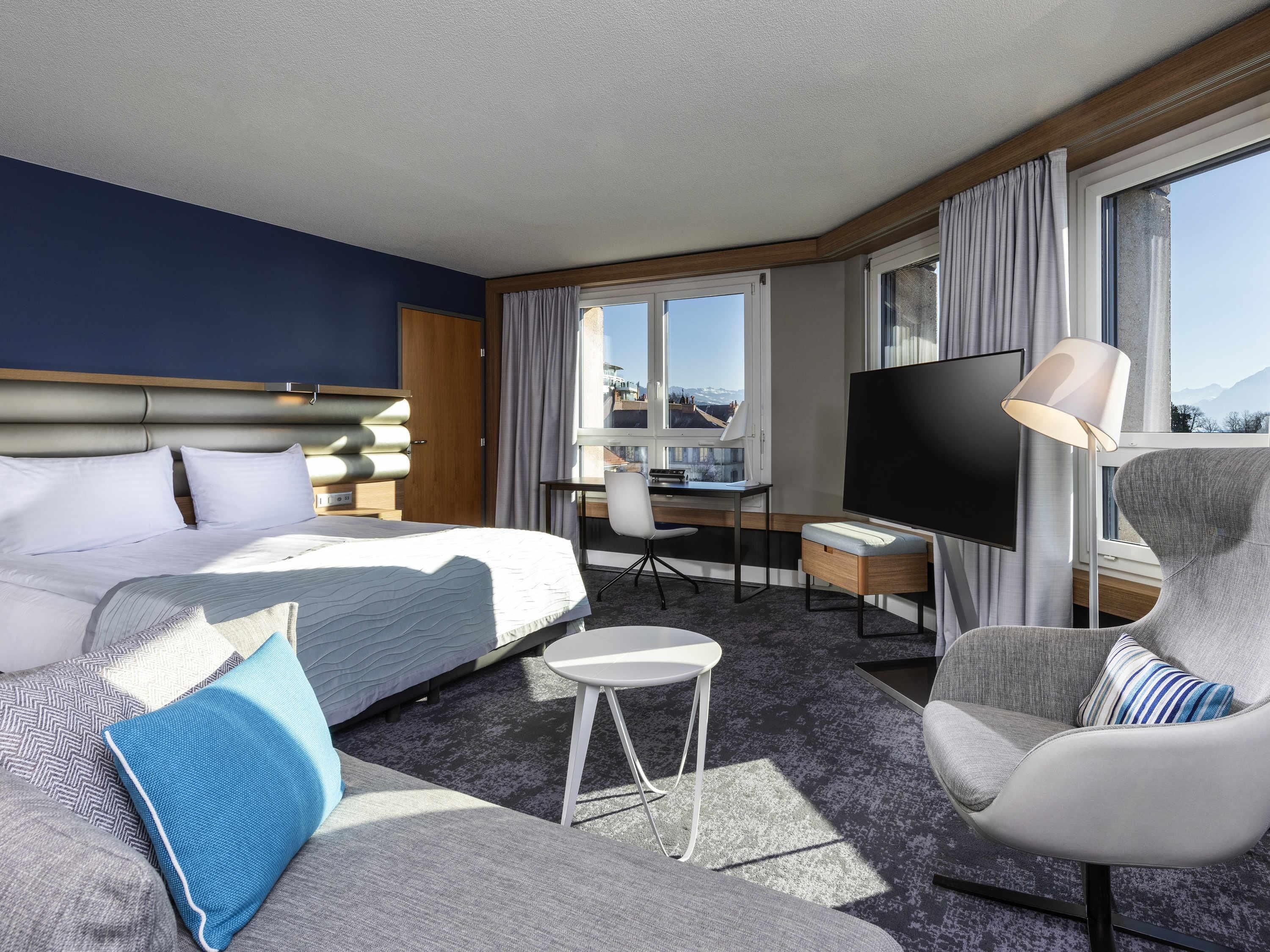 A hotel room with a bed, a desk, a chair, a TV, and large windows.