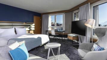 A hotel room with a bed, a desk, a chair, a TV, and large windows.