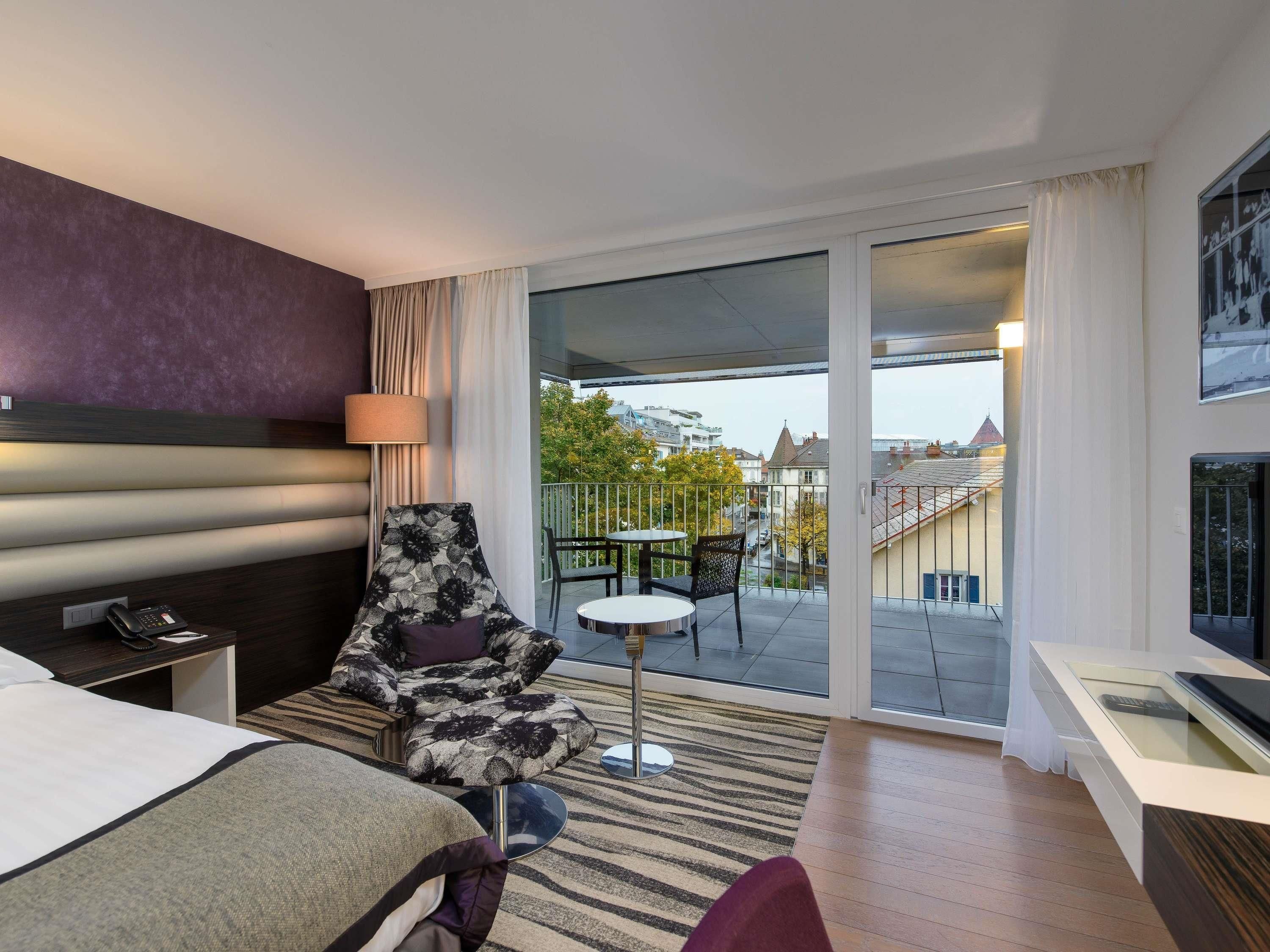 Ein Hotelzimmer mit Bett, Schreibtisch mit Telefon, Sessel, kleinen Tisch und Balkon mit Blick auf Gebäude und Bäume.