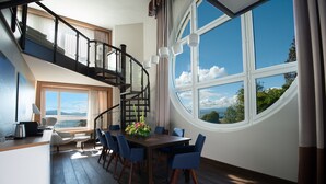 Un comedor con una mesa de madera y sillas azules, una escalera con barandillas negras, y ventanas amplias que ofrecen vistas al cielo y a los árboles.
